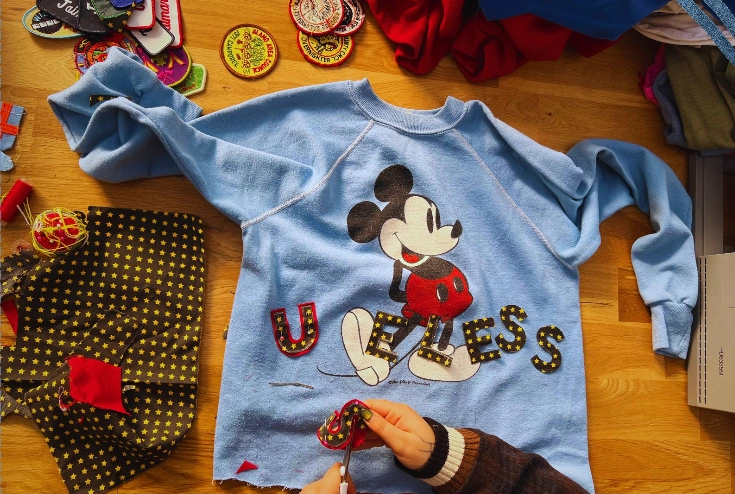 Sewing star-print USELESS letters onto a vintage blue Mickey Mouse sweatshirt, surrounded by patches and fabric